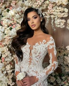 a woman wearing a wedding dress with long sleeves and flowers on the wall behind her