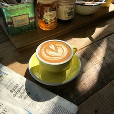 a cappuccino sits on a saucer next to some honey jars and a newspaper