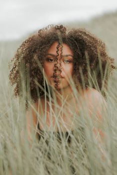 a woman with curly hair is standing in tall grass and has her face partially obscured by the grass