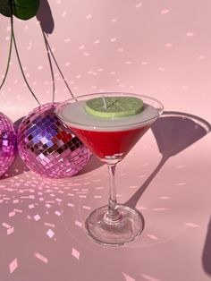 a martini with a lime slice on the rim next to two disco ball ornaments and a pink background