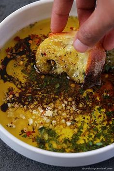 a person dipping food into a bowl of soup with bread and herbs on the side