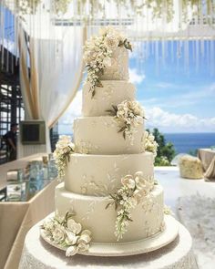 a white wedding cake with flowers on it sitting in front of an ocean view table