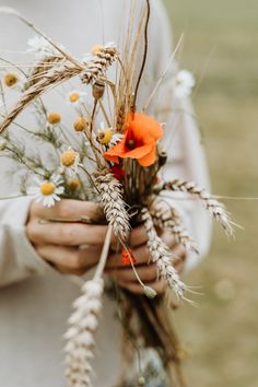 a woman holding some flowers and wheat stalks in her hands with an orange flower on top