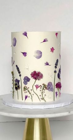 a white and purple cake with flowers on the top is sitting on a gold plate