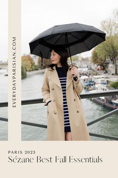 a woman in a trench coat holding an umbrella over her head and looking at the water