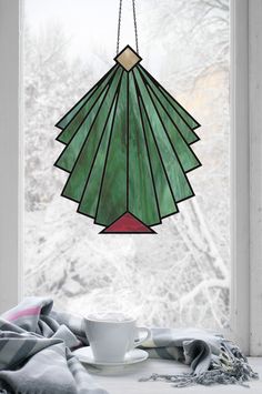 a cup and saucer sitting on a window sill in front of a stained glass christmas tree ornament