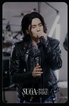 a man in a black leather jacket singing into a microphone with his hand to his mouth