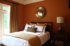 a bedroom with an orange wall and white bedspread, brown curtains, and a mirror on the wall