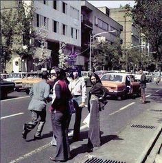 some people are standing on the side of the road in front of cars and buildings