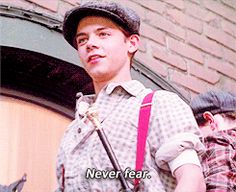 a young man wearing a hat and suspenders holding a microphone in front of a brick building