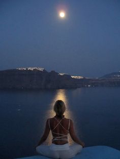 a woman is sitting on the edge of a cliff looking at the water and moon