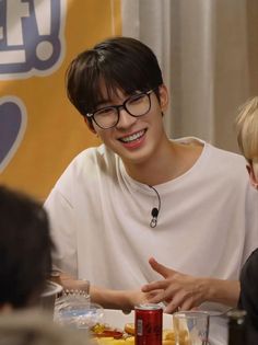 a young man with glasses sitting at a table talking to another person in front of him