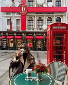 a woman sitting at a table drinking from a coffee cup in front of a red phone booth