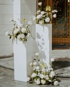 two tall white vases with flowers in them on the side of a street near a building