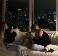 two women sitting on a couch with a dog in their lap looking out the window