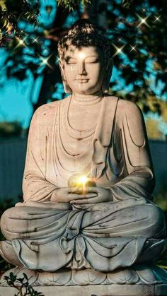 a buddha statue with a lit candle in its lap sitting under a tree on a sunny day