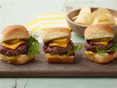 three hamburgers with cheese and lettuce on a cutting board next to chips