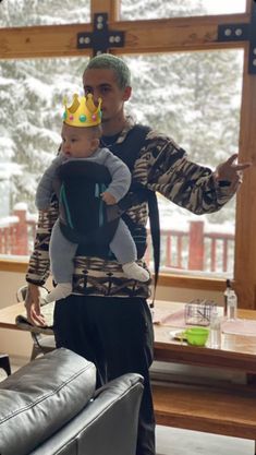 a man holding a baby wearing a crown on top of his head while standing next to a couch