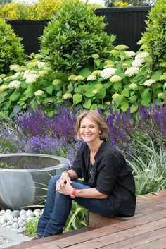 a woman sitting on a wooden bench in front of some bushes and flowers, smiling for the camera