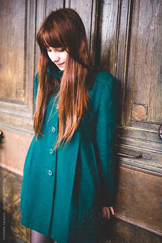 a woman with long red hair standing in front of a wooden door wearing a green coat