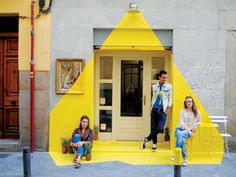 three people are sitting on the steps in front of a yellow building with a triangle painted on it