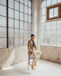 a woman sitting on top of a stool in an empty room with large windows behind her