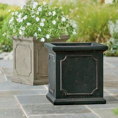 two black planters sitting on top of a stone floor next to white and green plants