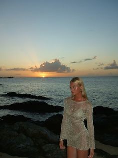 a woman standing on top of a rocky beach next to the ocean at sun set