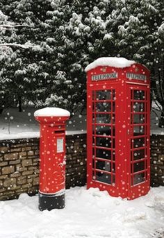 two red telephone booths covered in snow next to a brick wall and tree filled with snow