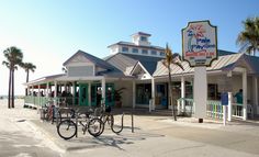 several bicycles are parked in front of the beachfront restaurant, with palm trees lining the sidewalk