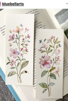 three cards with watercolor flowers on them sitting on top of an open book next to a plant