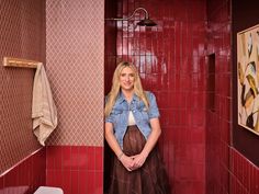 a woman standing in a bathroom next to a red tiled wall and toilet with a blue denim shirt on