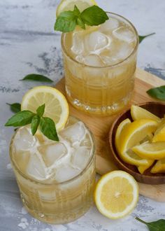 two glasses filled with lemonade and ice on top of a cutting board next to sliced lemons