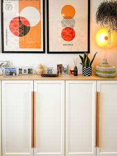 two framed pictures hang on the wall above a white cabinet with drawers and vases