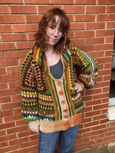 a woman standing in front of a brick wall wearing a multicolored sweater and jeans