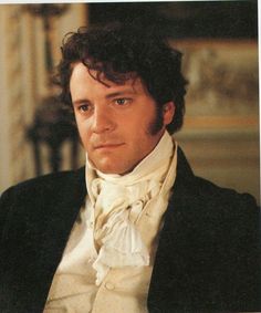 a man with curly hair wearing a black jacket and white shirt, sitting in a chair