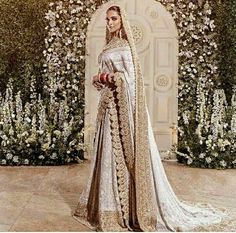 a woman in a white and gold bridal gown standing next to a flower covered wall