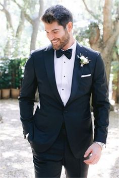 a man in a tuxedo smiles at the camera while standing under some trees