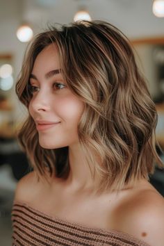 Woman with shoulder-length wavy hair smiling in a cozy, softly lit indoor space. Cinnamon Lowlights, Caramel Brown Hair, Neutral Skin Tone, Auburn Highlights