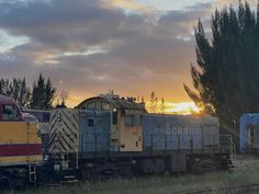 an old train is sitting on the tracks in front of some trees and buildings at sunset