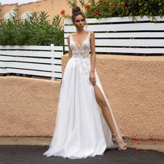 a woman wearing a white wedding dress with a slit in the side and flowers on the wall behind her