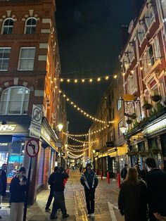 people are walking down the street in front of stores at night time with lights strung across the buildings