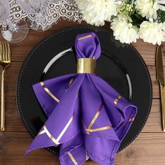 a black plate topped with purple napkins next to gold fork and knife on top of a wooden table