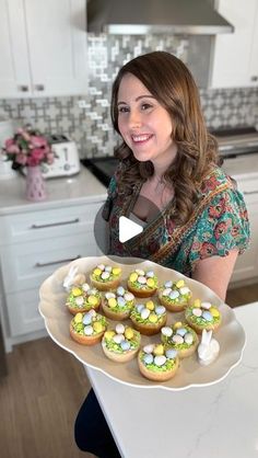 a woman holding a platter with cupcakes on it in the middle of a kitchen