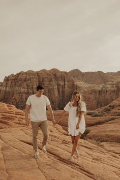 a man and woman holding hands while walking on rocks in front of the desert mountains