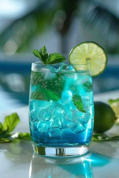 a glass filled with blue liquid and limes on top of a white tablecloth