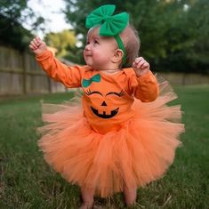 a baby girl dressed in an orange pumpkin outfit and green headband, standing on grass