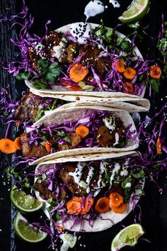 three tacos with meat, cabbage and carrots on a black surface next to lime wedges