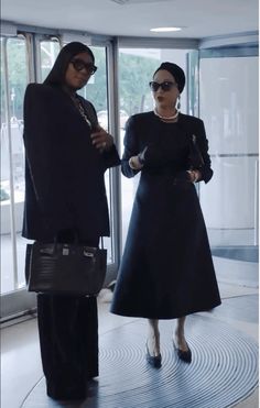 two women standing in front of large glass doors, one wearing a black dress and the other carrying a black handbag