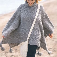 a woman walking on the beach wearing a gray sweater with tassels and a white bag